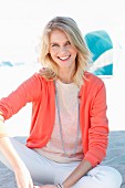 A blonde woman wearing a knitted jumper, a salmon-coloured cardigan and white trousers on the beach