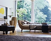 Cushions and blankets on couch next to black ottoman in front of panoramic window