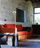 Orange sofa, side table and single allium flower in vase in front of rustic stone wall