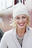 A blonde woman wearing a beige knitted jumper, a woollen coat and a white knitted hat