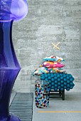 Murano-Glasgefäß vor buntem Kissenstapel und petrolfarbenem Wollkugel-Teppich vor Betonwand