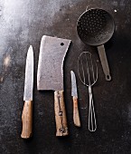 Alte Küchenwerkzeuge auf dunklem Untergrund: Fleischerbeil, Pfannenheber, Messer und Seiher