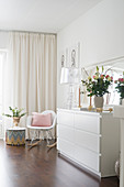 Vase of flowers and transparent table lamp on white chest of drawers, classic rocking hair and side table next to window with curtains