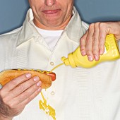 A man holding a hot dog in his hand and spilling mustard on his shirt