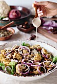 A woman is drizzling a bowl of roasted cauliflower salad with tahini dressing