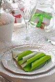 White place setting with green paper napkin folded into Christmas tree amongst artificial snow on table