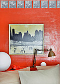Painting on red-painted wall with reading lamp on shelf at head of double bed