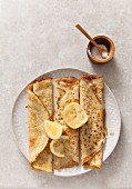 three rolled pancakes pn a off white texture plate with lemon wedges on a stone surface propped with wooden sugar bowl and spoon