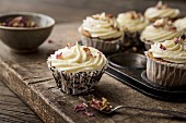 Rose and lemon cupcakes with rosewater and lemon frosting in a rustic baking tin decorated with dried edible rose petals with spoon and bowl of rose petals on a wooden board and table