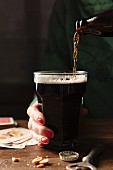 Bottle of Guinness being poured into a large glass being held by a hand with red nail varnish on a wooden table surrounds by the bottle top, bottle opener, nuts and playing cards