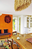 Holztisch mit Stühlen auf buntem Teppich und lila Sitzbank vor orangefarbener Wand