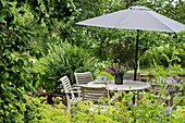 Round table, chairs and parasol on terrace in densely planted garden