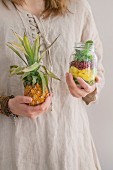 A woman in a linen dress holding a jar of fruit salad with mango, pomegranate seeds, lettuce and a fresh baby pineapple