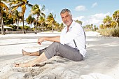 A man with grey hair wearing a white shirt and grey trousers sitting on the beach