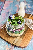 A quinoa salad with lambs lettuce, radicchio, rocket, croutons, goat's cheese and horned violets in a glass jar on a wooden board, with dressing in a glass bottle