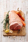 A raw joint of pork with an onion, carrots, rosemary and spices on a chopping board
