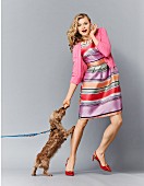 A blonde woman wearing a brightly striped dress, a pink cardigan and slingbacks with a dog