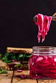 Pickled red onions are picked with a pair of tongs from a jar on a wooden board