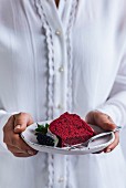 A woman with white shirt holding a slice of red velvet bundt cake with white glaze, a mulberry and mint
