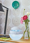 A ranunculus bouquet, an air humidifier and a lamp on a bedside table in front of patterned wallpaper