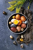 Brown and gray colored chicken and quail Easter eggs in black ceramic bowl with yolk, yellow flowers, sackcloth rag over black concrete texture background