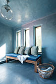 Holzbank mit blauen Kissen vor drei vertikalen Fenstern im blauen Raum