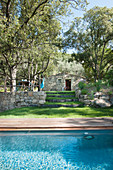 Swimming pool surrounded by wooden decking in Mediterranean garden with stone house in background
