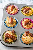 A metal muffin tray with savoury Frankfurter sausage muffins in colourful silicone cases. Crispy bacon, corn and green peas
