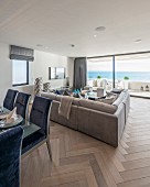 Blue upholstered dining chairs around table and pale sofa in front of glass wall in open-plan interior with sea view