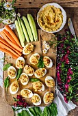 Hummus deviled eggs are served on a wooden board with cucumber and carrot sticks, arugula, and flowers