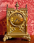 Ornate gilt table clock with Roman numerals against red brocade wallpaper
