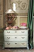 Pink glasses and table lamp on old chest of drawers against panelled wall