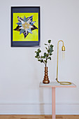 Vase of leaves and reading lamp on side table below colourful artwork