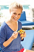 A blonde woman wearing a blue long-sleeved top and eating a fruit salad