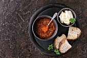 Bowl of red caviar with spoon served with sliced bread, butter and herbs on black wooden chopping board