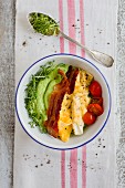 A breakfast bowl with avocado and cucumber cream, scrambled eggs, bacon and tomatoes