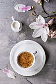 White cup of black coffee, served on white saucer with jug of cream and magnolia flower blossom branch
