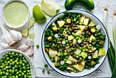 A bowl of potato salad with green lentils on a white background, Tahini dressing, peas, green apple slices, garlic and cucumber accompany