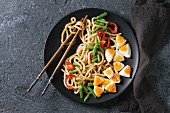 Cooking stir fry udon noodles, green beans, sliced paprika, boiled eggs, soy sauce with sesame seeds in black plate with wood chopsticks