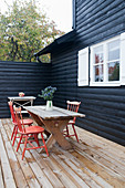 Red spoke-back chairs at wooden table on terrace of black wooden house