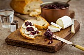 Copper pan filled with red onion chutney on a dark wooden chopping board with crusty bread vintage cutlery and sliced goat s cheese topped with chutney