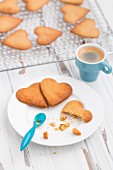 Cooling rack stacked with a grid of heart shaped biscuits on an aged white background with espresso and broken biscuits on a white plate with a blue espresso spoon