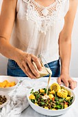 A woman wearing a white summer top and jeans pouring tahini dressing over lentil salad with sweetcorn and peaches