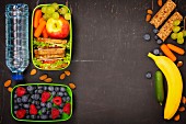 Sandwich, apple, grape, carrot, berry in plastic lunch box and bottle of water on black chalkboard