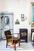 Antique wardrobe with mirror on door, log burner and armchair with green upholstery in living room with white wooden floor