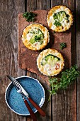 Baked homemade quiche pie in mini metal forms served with fresh greens, plate and cutlery on terracotta board