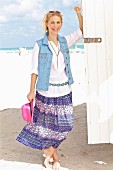 A young blonde woman on a beach wearing a white blouse, a denim gilet and a purple summer skirt