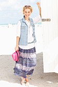 A young blonde woman on a beach wearing a white blouse, a denim gilet and a purple summer skirt