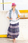A young blonde woman on a beach wearing a pink hat, a white blouse, a denim gilet and a purple summer skirt