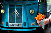 A hand holding a cone of sweet potatoes and chips in front of a food truck